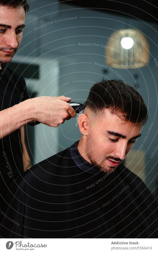 Junger Friseur, der einem Kunden mit einer elektrischen Haarschneidemaschine die Haare schneidet. Schermaschine Behaarung entgittern geschnitten jung Mann Salon
