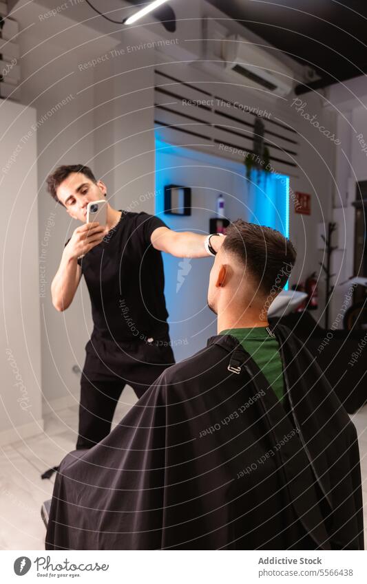 Friseur, der die neue Frisur eines Kunden im beleuchteten Salon festhält Foto Klient schwarz T-Shirt Mann Kaukasier Beleuchtung Zeitgenosse Telefon Fotokamera