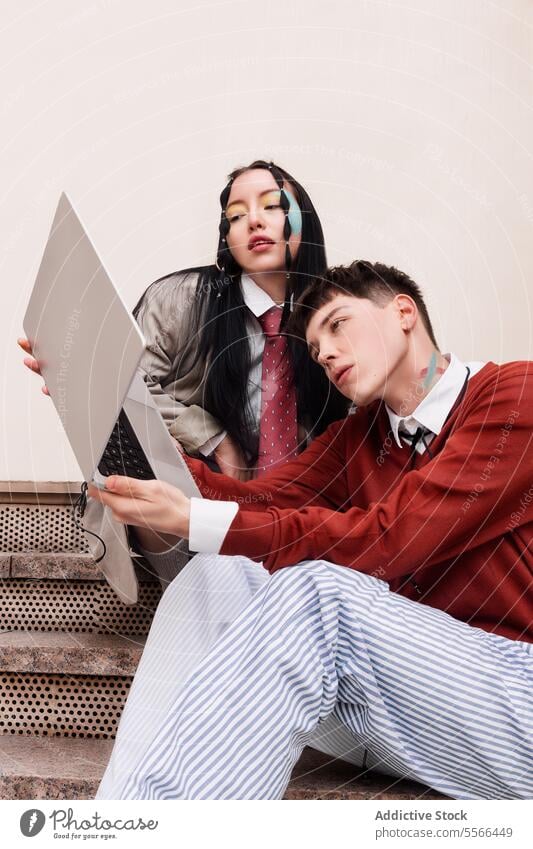 Zwei junge Modemodelle sitzen auf einer städtischen Treppe und schauen auf einen Laptop. Konzept der Arbeit. Gen-Z Frau Mann Make-up Krawatte Hemd rot Auge