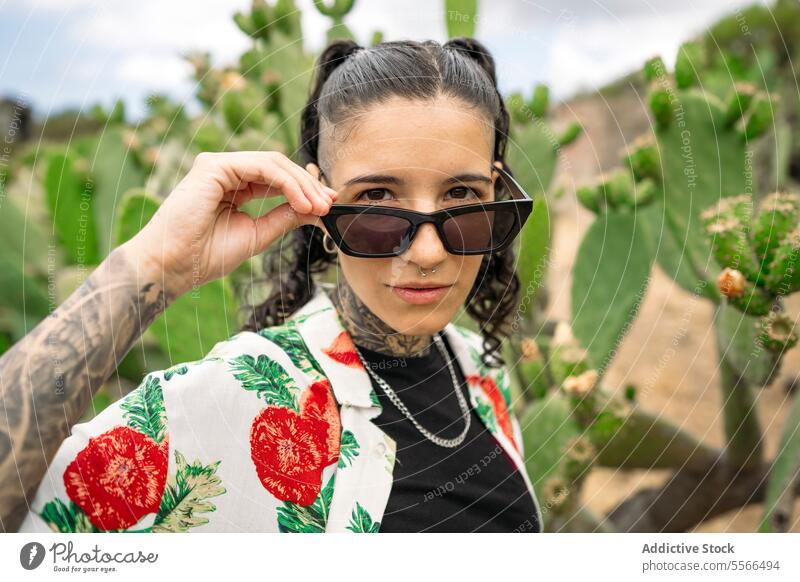 Tätowierte Frau, die auf einem unscharfen Kaktus steht und ihre Sonnenbrille verstellt stylisch Natur Tattoo Stehen attraktiv jung Mode hübsch Stil cool