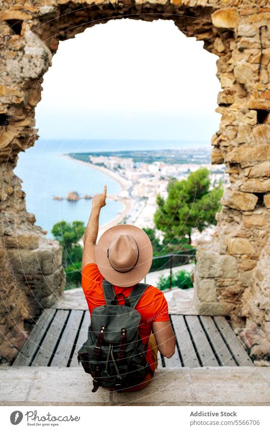 Ein Reisender gestikuliert vom historischen Bogen in Barcelona in Richtung Küstenstadt. Stein Mann Stadt Punkt Tourist orange vertikal Feiertage Hemd Hut