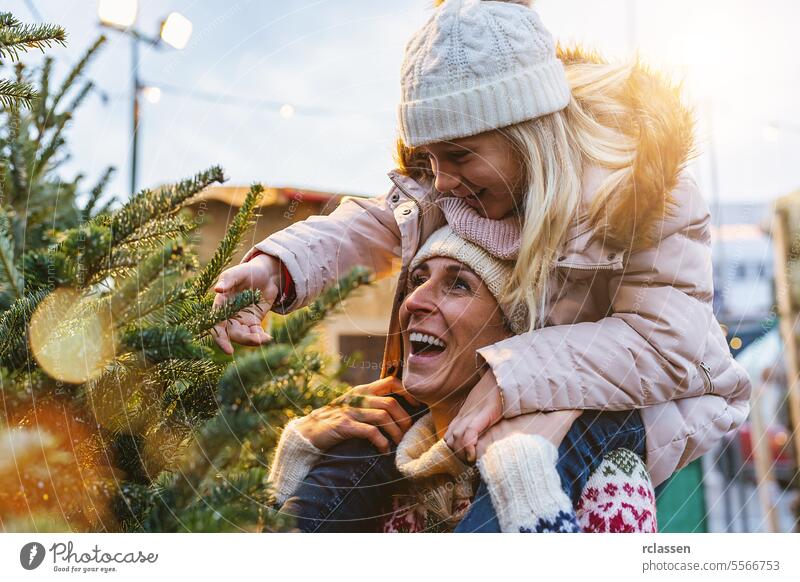 glückliche Mutter trägt ihre kleine Tochter Huckepack und sucht sich einen Weihnachtsbaum auf dem Straßenmarkt aus Kind überrascht Familie Glück skeptisch