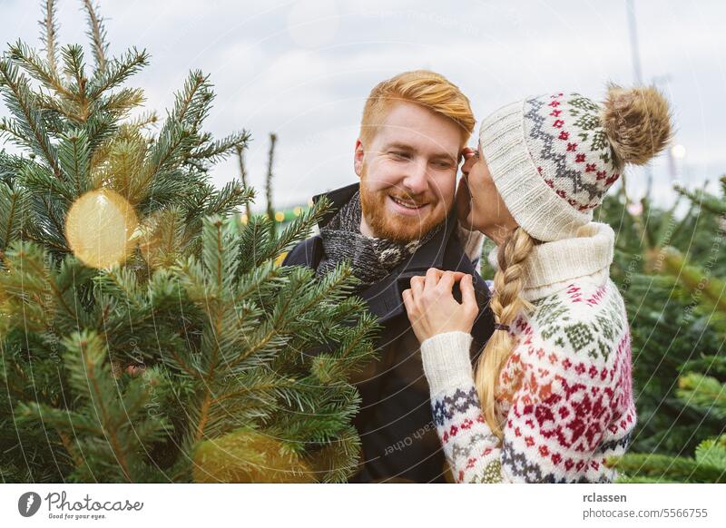 Paar sucht gemeinsam auf einem Markt einen Weihnachtsbaum aus, sie flüstert ihm etwas ins Ohr überrascht Familie Glück Rotschopf Tradition Wahl Lächeln Mann