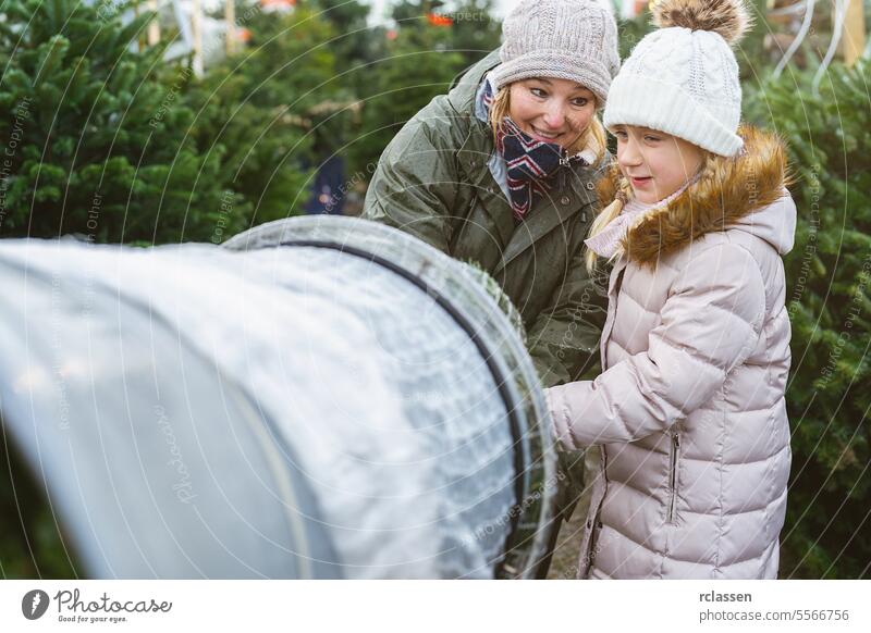 Glückliche Verkäuferin hilft einem Kind beim Einpacken eines gefällten Weihnachtsbaums, der in einem Plastiknetz auf einem Weihnachtsmarkt verpackt ist, Teamwork-Konzeptbild