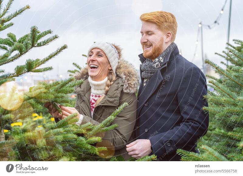 glückliches Paar sucht Weihnachtsbaum auf dem Straßenmarkt aus überrascht Familie Glück skeptisch Rotschopf Tradition Wahl Lächeln Mann Liebespaar Kauf