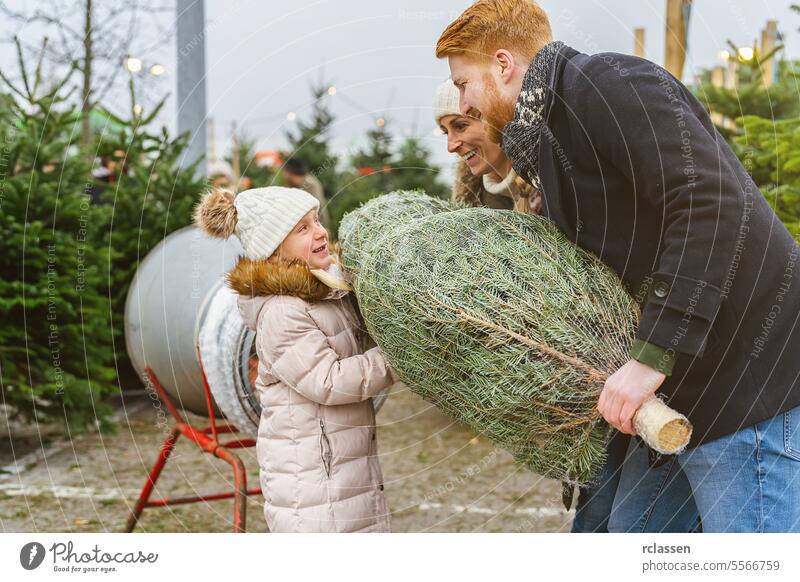 Familie, die auf einem Weihnachtsmarkt einen gefällten, in ein Plastiknetz verpackten Weihnachtsbaum einwickelt Teamwork kaufen Hilfsbereitschaft Kind Tochter