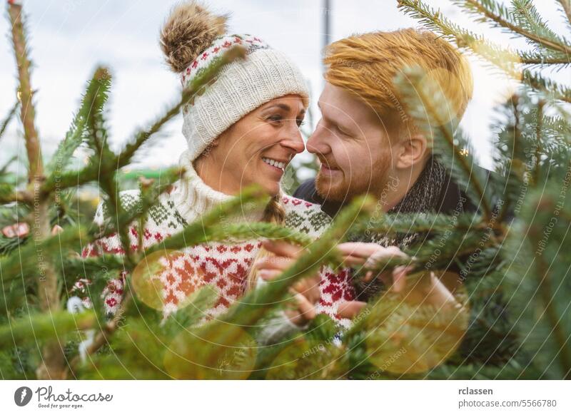Verliebtes Paar kauft Weihnachtsbaum auf dem Straßenmarkt betrachten Kuss Familie Glück Rotschopf Tradition Wahl Lächeln Mann Liebespaar Kauf Bauernhof Kiefer
