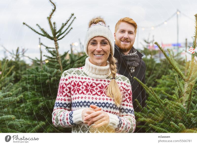 Glückliches Paar auf einem Markt will einen Weihnachtsbaum kaufen und schaut in die Kamera Blick überrascht Familie Rotschopf Tradition Wahl Lächeln Mann