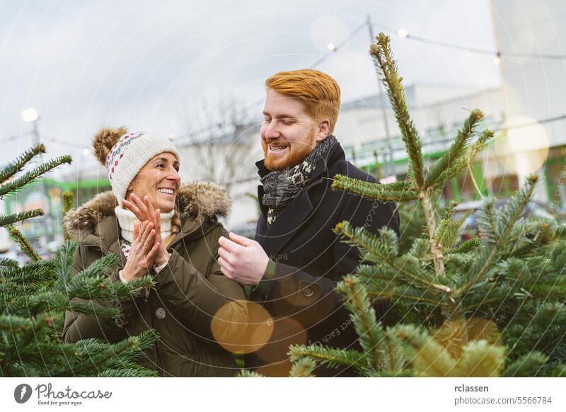 Glückliches Paar sucht gemeinsam einen Weihnachtsbaum auf einem Markt aus überrascht Familie skeptisch Rotschopf Tradition Wahl Lächeln Mann Liebespaar Kauf