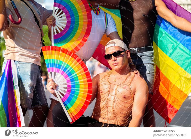 Selbstbewusster Mann mit Papierfächer bei LGBT-Pride-Veranstaltung Sonnenbrille gold Halskette Pose Stolz Ventilator Fahne Sonnenlicht pulsierend Farbe