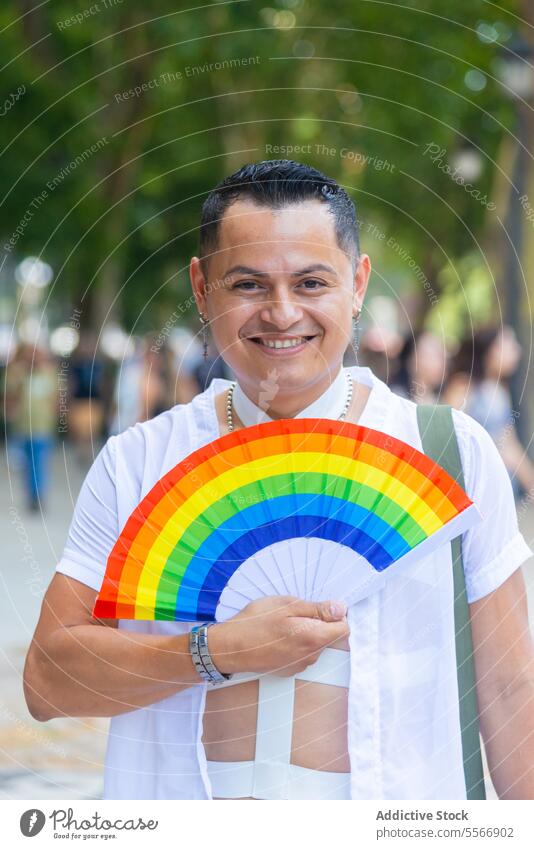 Glücklicher erwachsener Mann mit einem LGBT-Stolz-Fan heiter Menge urban Ventilator weiß Hemd Ohrringe Lächeln Freude Feier lgbtq pulsierend Farbe im Freien