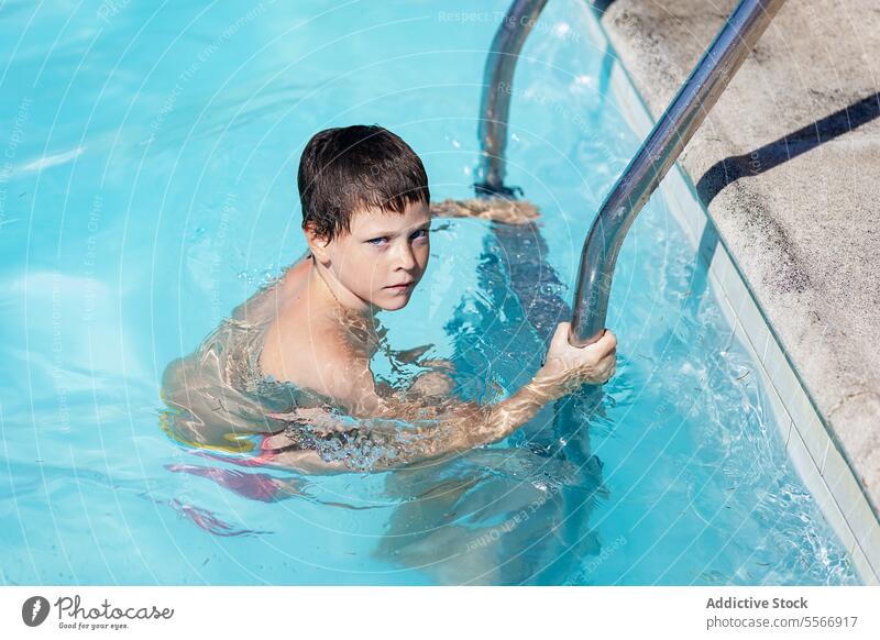 Junge in einem Schwimmbecken, der sich mit aufmerksamem Blick an einer Leiter festhält. Pool Laufmasche Wasser Starrer Blick Sommer schwimmen blau Kind Porträt