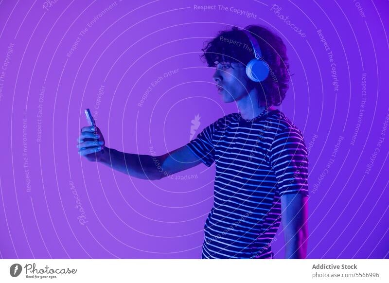 Lateinamerikanischer Mann mit Kopfhörern, der sein Smartphone lila beleuchtet lockig Behaarung gestreift Hemd purpur Starrer Blick Licht Ansicht Bildschirm