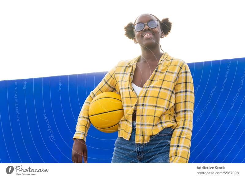 Glückliche nachdenkliche Frau hält Basketball während eines sonnigen Tages Lächeln Brille Wegsehen Sonnenlicht Stehen lässig Wand blau gelb Plaid Hemd Sport
