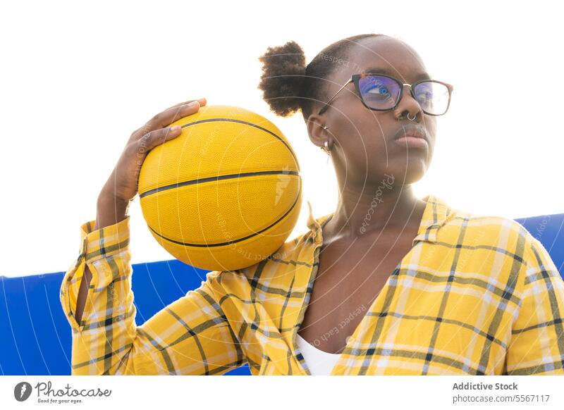 Nachdenkliche Frau hält Basketball an einem sonnigen Tag nachdenklich ernst Brille Wegsehen Sonnenlicht lässig gelb Plaid Hemd Sport Freizeit Wochenende