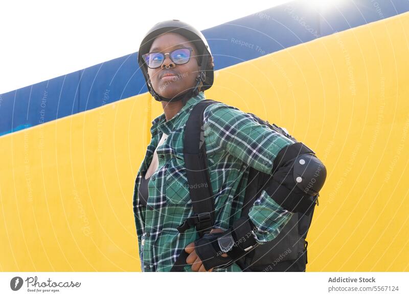 Nachdenkliche Frau mit Helm und Brille auf dem Skatepark nachdenklich Schutzhelm Wegsehen Sonnenlicht Ausrüstung Rucksack Stehen lässig Wand gelb Plaid Hemd