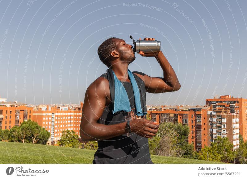 Mann trinkt nach dem Sport im Freien trinken Flasche rostfrei Stahl Stadtbild weiß Hydratation Grün Baum Skyline Park aktualisieren Hals Handtuch Getränk Wasser