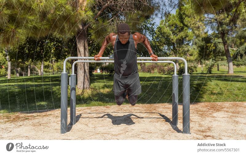 Schwarzer Mann in Sportkleidung führt eine Parallelbarren-Dip-Übung in einem Park mit üppigen Bäumen im Hintergrund aus parallel Bar sportlich im Freien Stärke