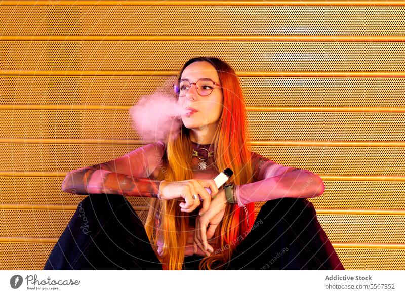 Junge Frau atmet vor orangefarbenem Hintergrund aus Steigung Behaarung Brille purpur durchlöchert E-Zigarette beschaulich Pose Stil Mode sitzend