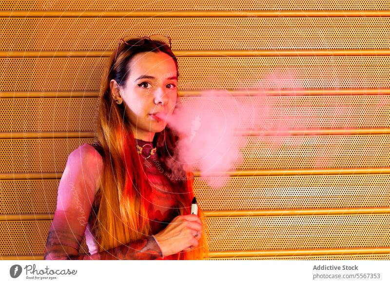 Junge Frau atmet vor orangefarbenem Hintergrund aus Dampf ausatmen texturiert pulsierend Behaarung Stil Kleidung selbstbewusst jung Mode Rauch E-Zigarette