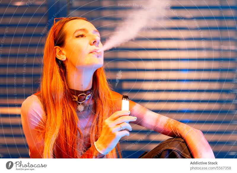 Junge Frau atmet in der Nähe eines blauen Metallhintergrunds aus Behaarung vaping E-Zigarette metallisch Hintergrund orange Illumination Ambiente Licht