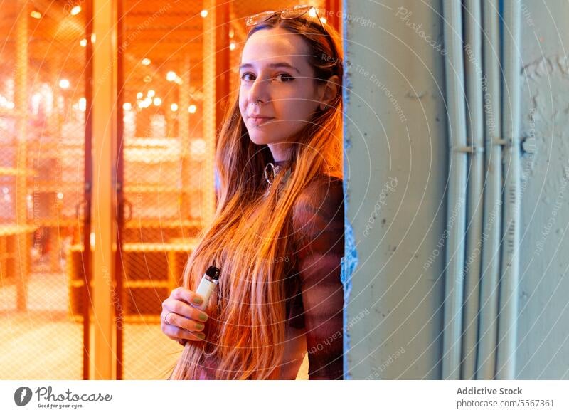 Junge Frau mit E-Zigarette vor heiterer städtischer Kulisse Behaarung orange ineinander greifen Zaun blaugrün Wand urban Gelassenheit Hintergrund Metall Röhren