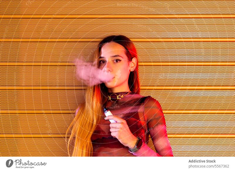 Junge Frau atmet vor orangefarbenem Hintergrund aus Dampf ausatmen texturiert pulsierend Behaarung Stil Kleidung selbstbewusst jung Mode Rauch E-Zigarette