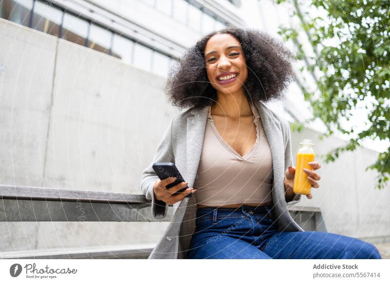Ethnische Frau mit Smartphone und Saft auf der Straße lockig Behaarung im Freien modern Gebäude Hand Flasche trinken Browsen sitzen urban Erfrischung Pause