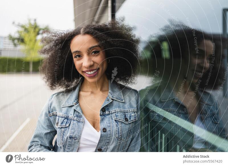 Ethnische junge Frau, die lächelt und sich gegen eine spiegelnde Glasscheibe lehnt lockig Behaarung Jeansstoff Jacke weiß Top Lächeln Lehnen