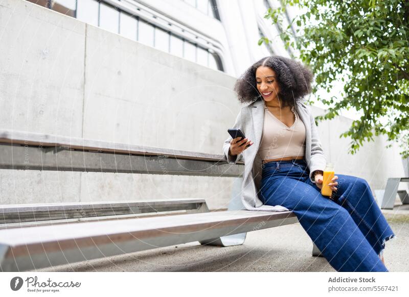Ethnische Frau mit Smartphone und Saft auf der Straße lockig Behaarung im Freien modern Gebäude Hand Flasche trinken lesen Browsen Nachricht sitzen urban