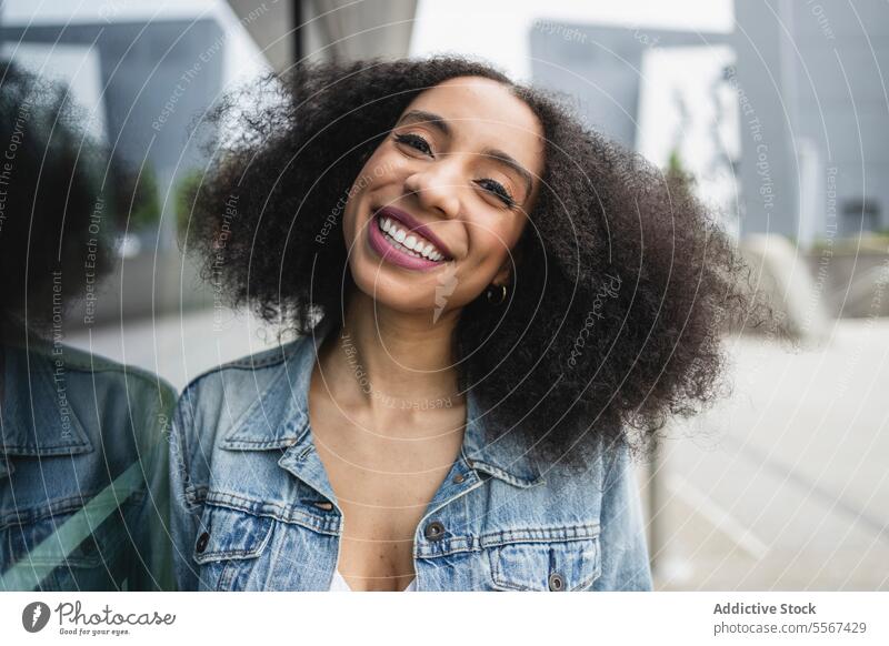 Ethnische junge Frau, die lächelt und sich gegen eine spiegelnde Glasscheibe lehnt lockig Behaarung Jeansstoff Jacke weiß Top Lächeln Lehnen