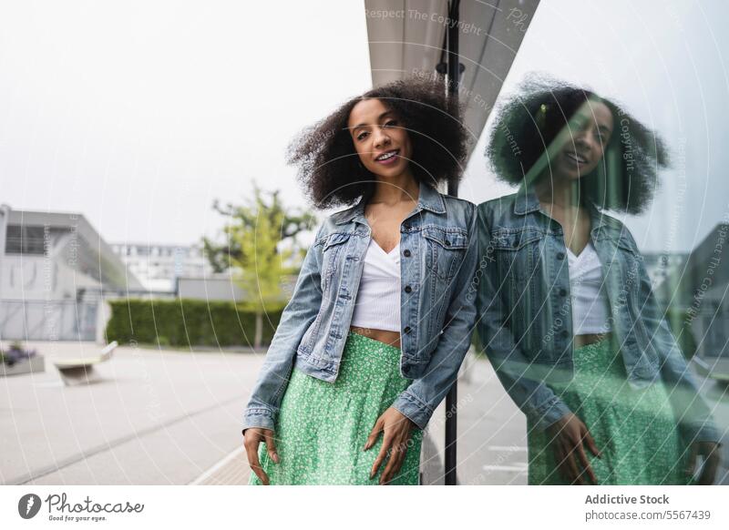 Ethnische junge Frau, die lächelt und sich gegen eine spiegelnde Glasscheibe lehnt lockig Behaarung Jeansstoff Jacke weiß Top grün Rock Lächeln Lehnen