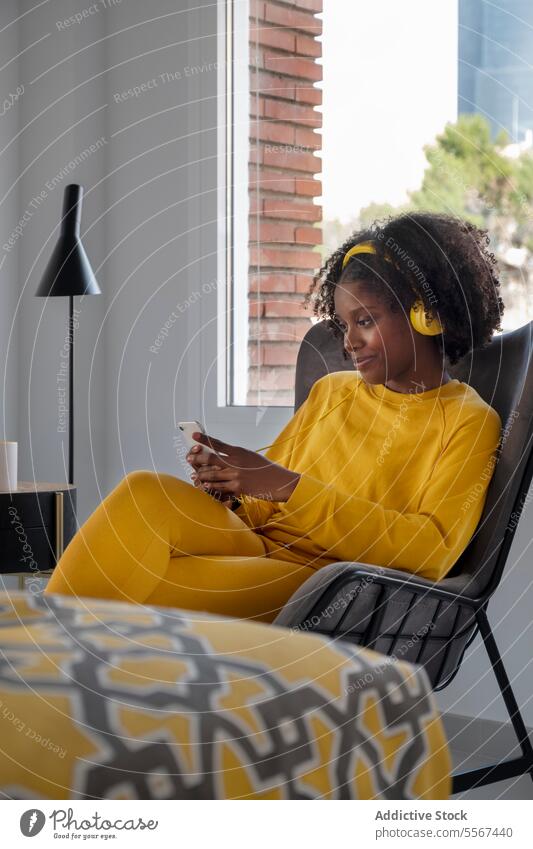 Schwarze Frau lebt entspannt in der Stadt gelb Outfit Appartement Musik Kopfhörer Afroamerikaner Telefon Browsen modern Innenbereich Fenster Stuhl ethnisch