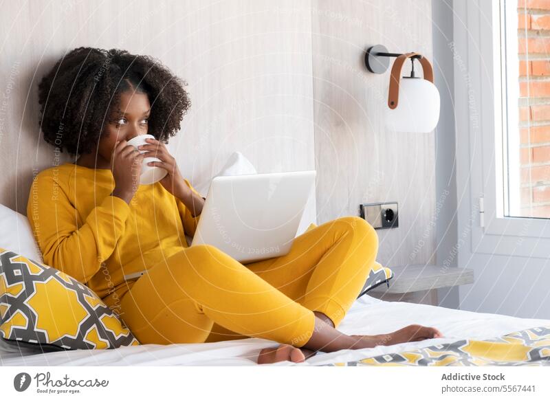 Schwarze Frau in gemütlicher Arbeitssituation zu Hause gelb Kaffee Laptop Bett heimwärts Innenbereich modern Fokus Kissen Wand Lampe Fenster Baustein