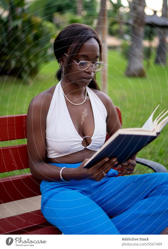 Afroamerikanische Frau liest im Garten Afroamerikaner lesen Buch Fokus Brille Bank im Freien grün entspannt Halfter weiß blau Hose Bäume beschaulich fleißig