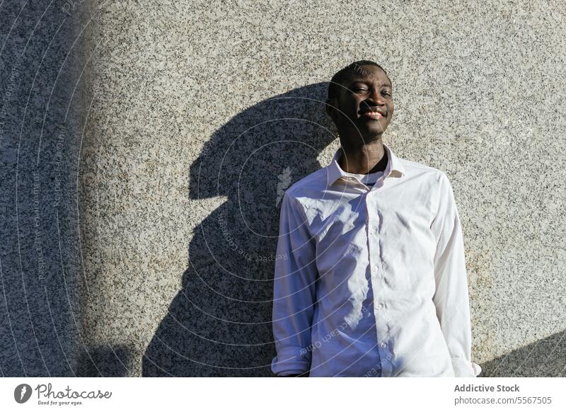 Lächelnder Mann im Sonnenlicht Schatten Textur Wand Hemd Gesicht Freude Lehnen urban im Freien weiß Inhalt Gelassenheit Pose Glück männlich Profil Sommer Jugend