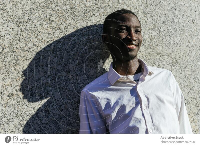 Lächelnder Mann im Sonnenlicht Schatten Textur Wand Hemd Gesicht Freude Lehnen urban im Freien weiß Inhalt Gelassenheit Pose Glück männlich Profil Sommer Jugend