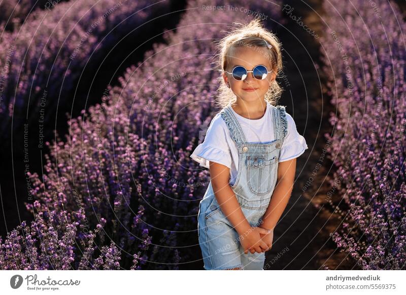 adorable Kind Mädchen in Lavendelfeld auf Sonnenuntergang. lächelnd Kind in Sonnenbrille, Jeans Overall hat Spaß an der Natur am Sommertag. Familientag, Urlaub, holiday.International Children's Day,
