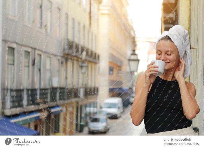 Junge schöne Frau in einem Handtuch auf dem Kopf trinkt Kaffee oder Tee und lächelnd niedlich stehend auf Balkon auf ueropean Straße Hintergrund. lifestyle Morgen. Konzept der inländischen oder Reisen Lebensstil.