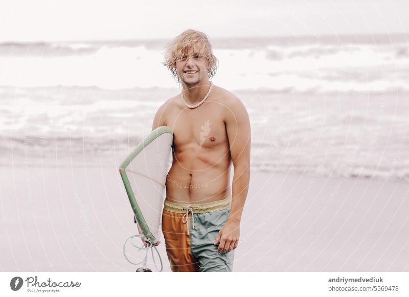 Handsome fit jungen blonden Mann mit Mock-up-Surfbrett wartet auf Welle zum Surfen vor Ort am Meer Ozean Strand mit schwarzem Sand und schaut in die Kamera. Konzept der Sport, Fitness, Freiheit, Glück, neue moderne Leben