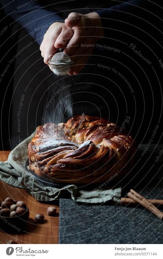 Frisch gebackenes Zimtbrot, mit Menschenhänden, die Puderzucker auf die Oberfläche streuen, in einer stimmungsvollen Umgebung in der Küche Aktion