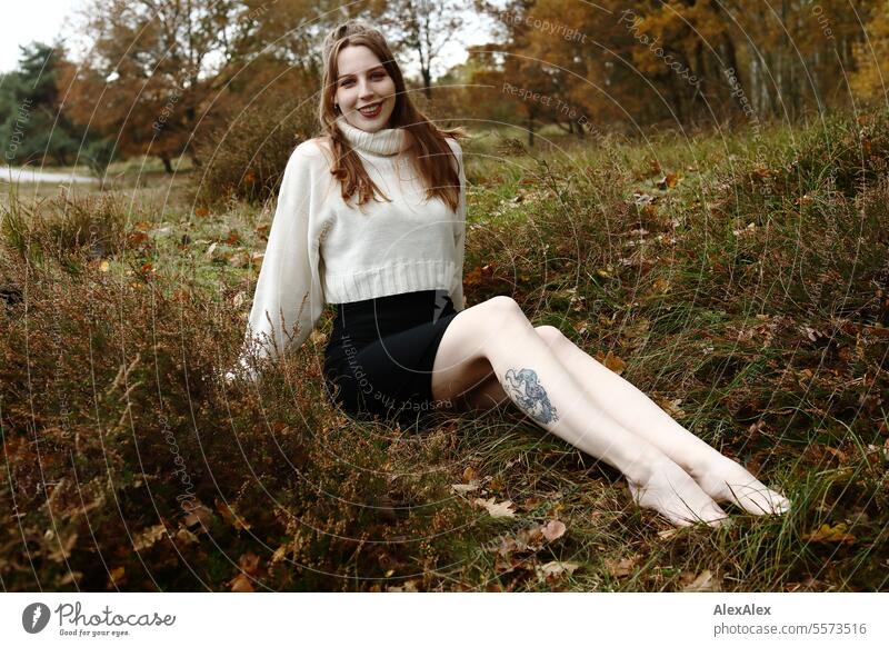 Junge Frau in weißem Wollpullover und schwarzem Rock sitzt barfuß in einer norddeutschen Heidelandschaft im Herbst im Gras zwischen Heidekraut und lächelt