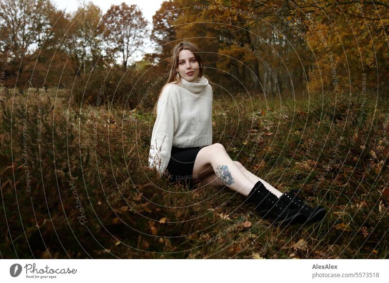 Junge Frau in weißem Wollpullover und schwarzem Rock sitzt in einer norddeutschen Heidelandschaft im Herbst im Gras zwischen Heidekraut junge Frau groß