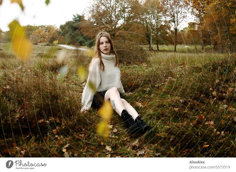 Junge Frau in weißem Wollpullover und schwarzem Rock sitzt in einer norddeutschen Heidelandschaft im Herbst im Gras zwischen Heidekraut junge Frau groß
