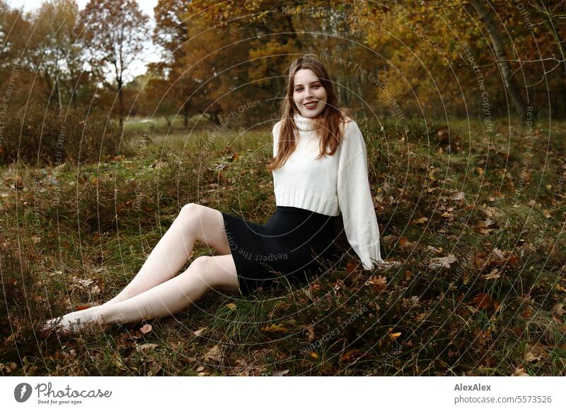 Junge Frau in weißem Wollpullover und schwarzem Rock sitzt barfuß in einer norddeutschen Heidelandschaft im Herbst im Gras zwischen Heidekraut und lächelt