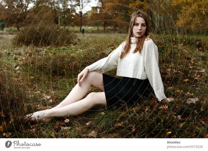 Junge Frau in weißem Wollpullover und schwarzem Rock sitzt barfuß in einer norddeutschen Heidelandschaft im Herbst im Gras zwischen Heidekraut junge Frau groß