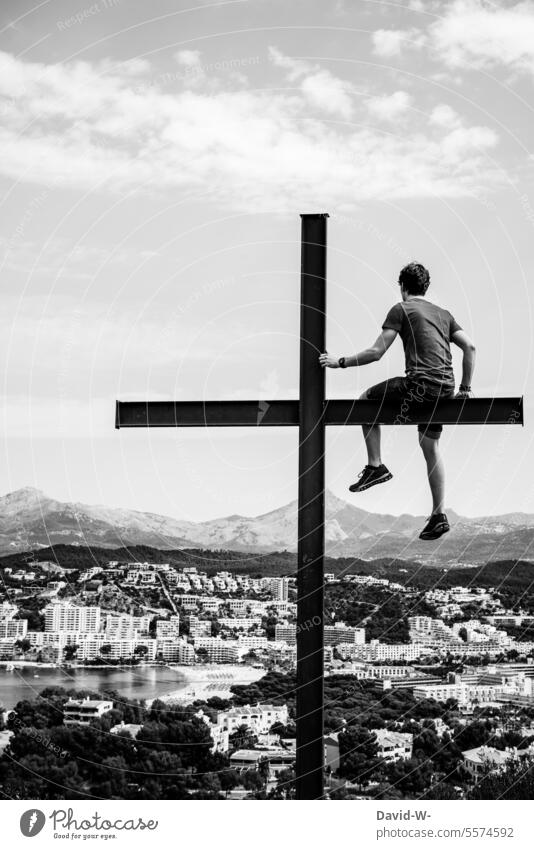 Junger Mann sitzt auf einem Kreuz und schaut in die Ferne Glaube Gott Religion Hoffnung Spiritualität Willenskraft Symbole & Metaphern Stadt Berg Rückansicht