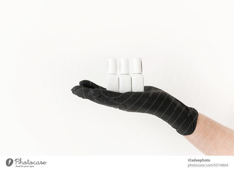 Weiße Tiegel mit Nagellack auf der Handfläche in einem schwarzen Nitrilhandschuh auf weißem Hintergrund. Maniküre Glas nageln Firnis polnisch Handschuh Frau