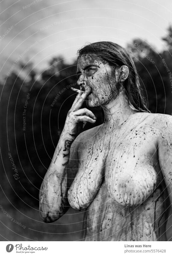 Ein Schwarz-Weiß-Bild mit einem nackten und wilden Mädchen darin. Sie raucht eine Zigarette und zeigt ihre nackten, sexy Kurven, mit etwas Schlamm und Körperfarbe auf ihr. Ein nacktes Model fühlt sich wild und frei in ihrer eigenen Haut. Und ein bisschen teuflisch.