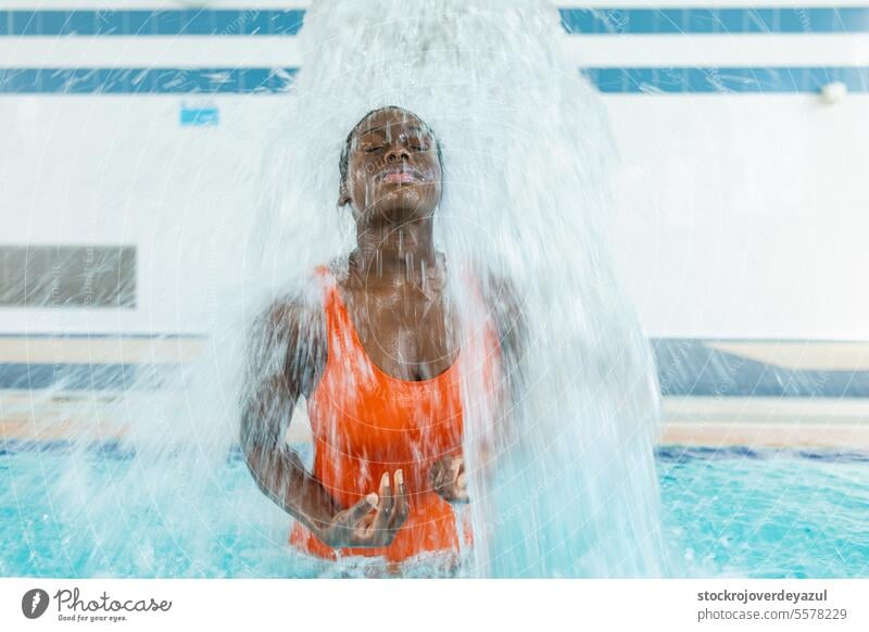 Schwarze junge Frau massiert ihren Rücken unter einem Wasserstrahl im Schwimmbad schwarz Pool Person Sport blau schwimmen aktiv erhitzt Badeanzug orange